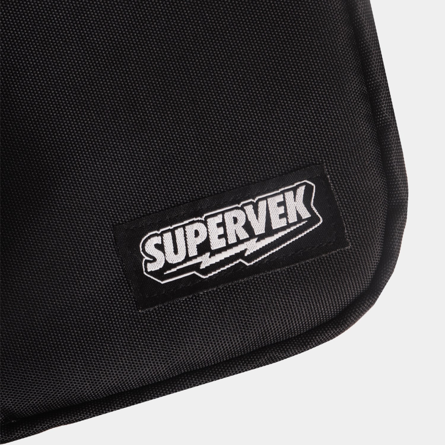 Detailed close-up of stitched Supervek logo patch on black "Sling" bag with textured fabric and Classic Black #color_Classic Black